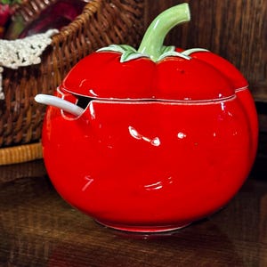 Puede incluir: Un recipiente de cerámica roja brillante con forma de tomate, con tallo y hojas verdes. Una pequeña cuchara blanca descansa en el interior. La tapa está ligeramente entreabierta. Un artículo de cocina encantador.