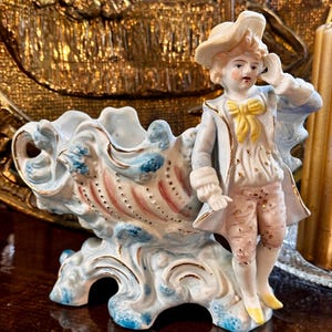 Puede incluir: Una figurita de porcelana de un niño con un traje azul y blanco con una faja amarilla, de pie sobre una base en forma de concha de mar azul y blanca con detalles dorados.