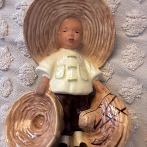 May include: A ceramic figurine of a young person wearing a large straw hat and white shirt with green accents. The figure is holding two baskets, one in each hand. The figurine is painted in shades of brown, white, and black.