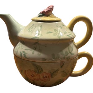 May include: A ceramic teapot with a floral design. The teapot has a lid with a pink flower on top. The teapot is a two-piece set, with a smaller cup attached to the bottom.
