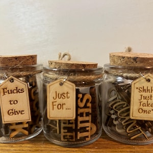 May include: Three glass jars with cork lids and wooden tags. The jars contain wooden letters spelling out phrases like "Fucks to Give", "Just For... SHIT", and "Shhh... Just Take One". The jars are tied with twine.