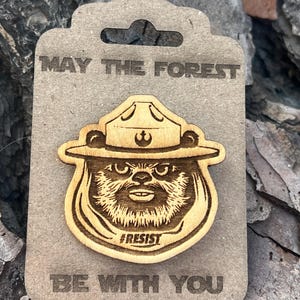 May include: Wooden pin featuring a bear wearing a ranger hat, with the text "MAY THE FOREST BE WITH YOU" and "#RESIST". The pin is attached to a cardboard backing. The design is laser-cut.