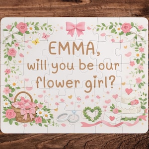 May include: A white puzzle with a floral design and the text "EMMA, will you be our flower girl?" The puzzle features pink roses, a basket of flowers, a cake, and a bow. The puzzle is on a wooden surface.