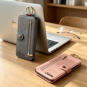 Peut inclure: Deux étuis de téléphone rectangulaires en cuir texturé avec une poche zippée verticale, une fermeture à bouton-pression et un anneau métallique doré sur le dessus. Un étui est gris avec le nom Charles, l'autre est rose avec le nom Chloe.