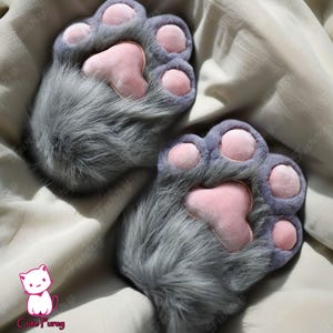 以下が含まれることがあります： ピンクの肉球が付いたグレーの猫の足の形をしたスリッパ。スリッパは柔らかく、ふわふわした生地でできており、毛皮の尻尾が付いています。