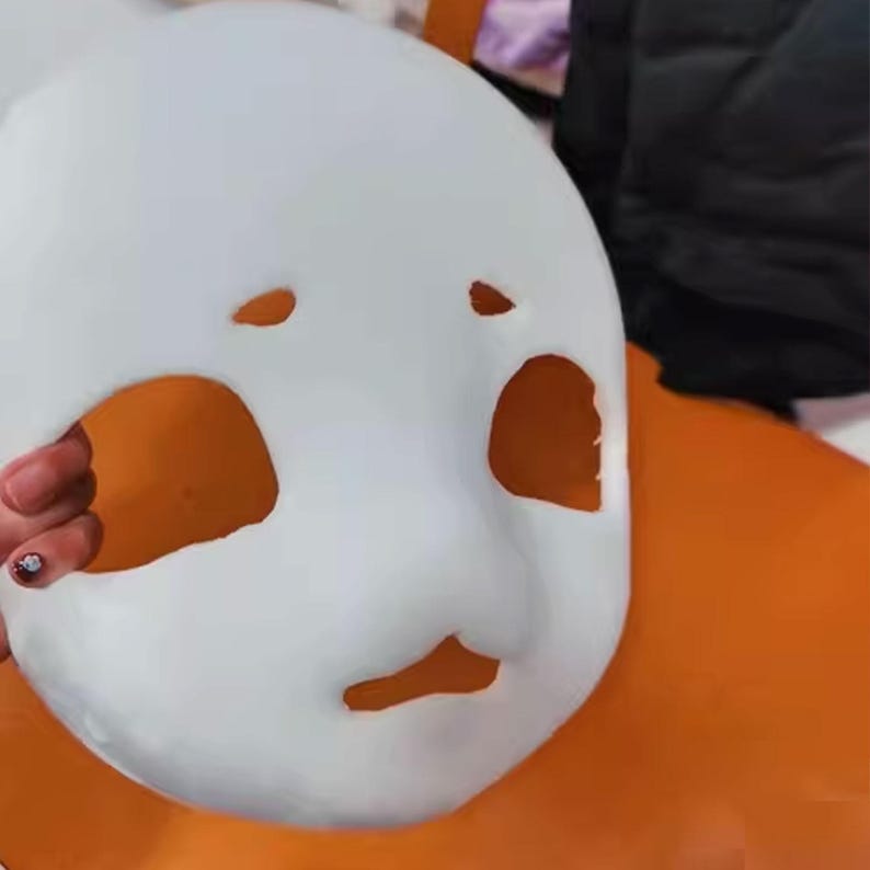 May include: A white mask with cut-out eye and mouth holes, and orange accents. The mask has a smooth surface and is held by a hand. The mask is likely for costume or decorative purposes.