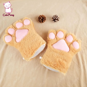 以下が含まれることがあります： ピンクの肉球が付いた茶色のぬいぐるみ製の猫の手あてが2つ。手あては薄い茶色の表面の上に置かれています。