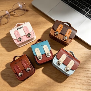 May include: Five small, rectangular, faux leather cases shaped like miniature satchels with top handles and dual snap-button straps. Each case features a unique colour combination and is personalised with the names Oliver, Noah, James, Henry, and Emma.