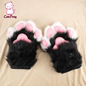 以下が含まれることがあります： ピンクの肉球が付いた黒と白の毛皮の手袋人形のペア。人形は手首に黒いゴムバンドが付いています。