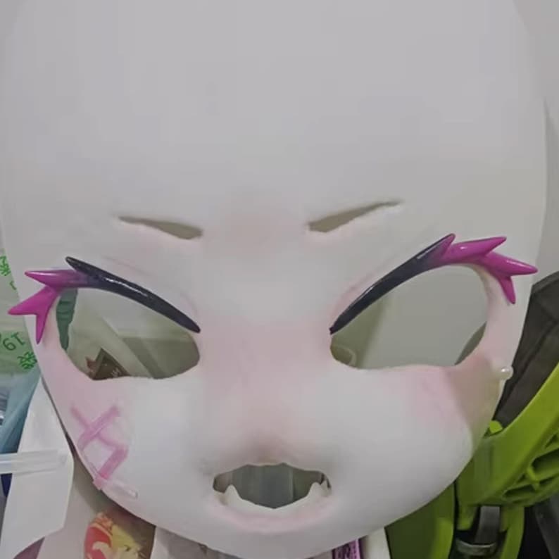 May include: A white mask with cut-out eye holes, a wide open mouth, and pink blush on the cheeks. The mask has black eyebrows with pink accents. A pink cross is on the left cheek.