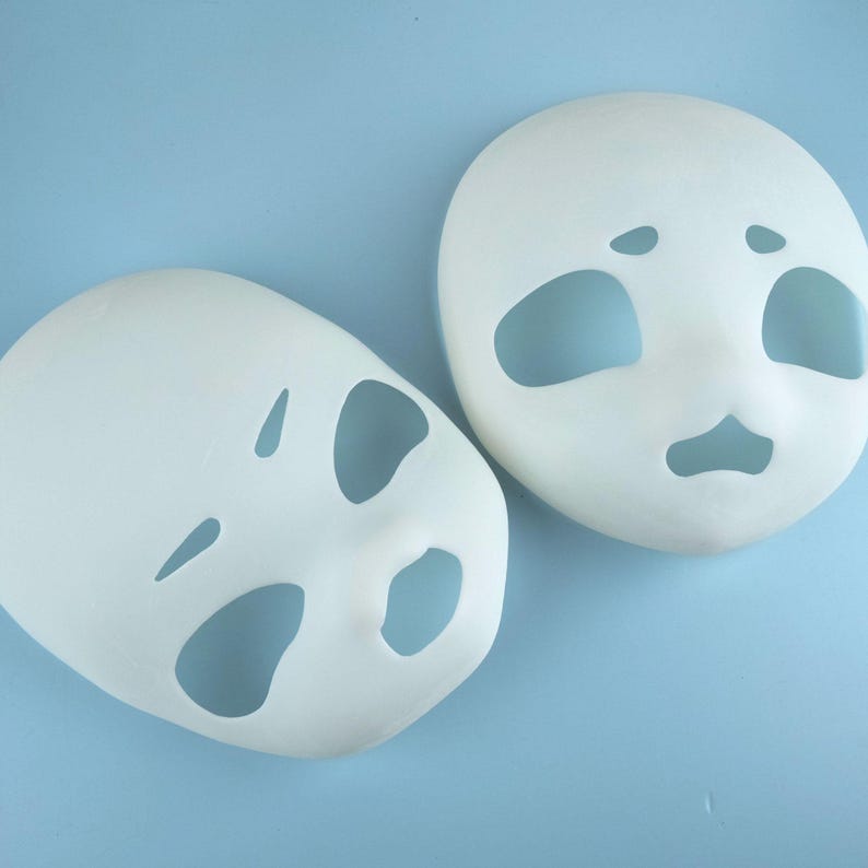 May include: Two white, blank face masks with eye, nose, and mouth cutouts. The masks are oval-shaped and sit on a light blue surface. The masks are ready to be painted or decorated.