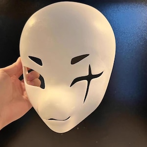 May include: A white mask with cut-out eye holes and a small mouth opening. Black markings on the right side of the mask resemble a cross. The mask is smooth and appears to be made of a solid material.