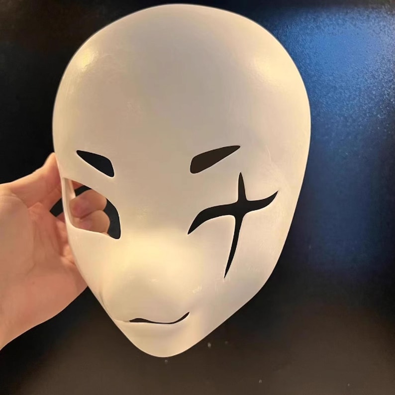 May include: A white mask with cut-out eye holes and a small mouth opening. Black markings on the right side of the mask resemble a cross. The mask is smooth and appears to be made of a solid material.