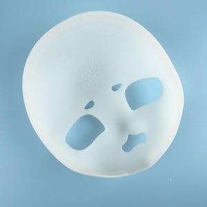May include: A white, face-shaped mask with eye, nose, and mouth cutouts. The mask has a smooth surface and a slightly rounded shape. A small embossed design is visible near the top. The background is a solid light blue.