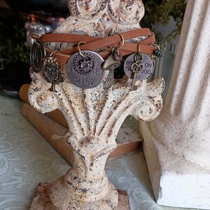 Puede incluir: Un brazalete de cuero marrón con múltiples encantos, incluyendo un árbol, un engranaje con el número 2167, un hongo, una llave y un encanto con el texto "Para el bien". El brazalete se muestra en un soporte de metal blanco.