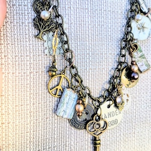 May include: A vintage-style necklace with multiple charms in shades of bronze and gold. Charms include a peace sign, a tree, a key, and a tag that reads "Lander 839". The necklace has a layered chain design and features small beads and a pearl.