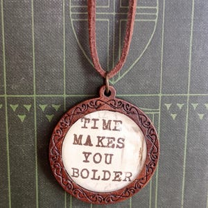May include: A brown wooden pendant necklace with a white background and the text "TIME MAKES YOU BOLDER" in black lettering.