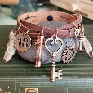 Puede incluir: Un brazalete de cuero marrón con varios encantos, incluyendo una llave en forma de corazón, una pequeña botella, un engranaje, un encanto con el número 18 y un pequeño objeto de metal. El brazalete está apoyado en una piedra gris.