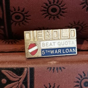 May include: A rectangular Diebold pin with the text "DIEBOLD" at the top. Below, it reads "BEAT QUOTA" and "5TH WAR LOAN" on a blue background. The pin features a red and white emblem. The background is a patterned, reddish-brown surface.
