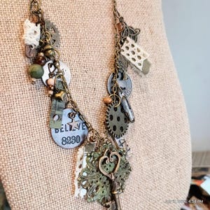 Puede incluir: Un collar artesanal con estética steampunk. Presenta una variedad de colgantes, incluyendo engranajes, llaves y una etiqueta que dice "BELIEVE 8230". La paleta de colores incluye acentos en bronce, verde y crema, creando un accesorio único y artístico.