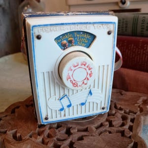 Puede incluir: Caja de música de juguete Fisher-Price vintage. La caja blanca y azul presenta un dial con "TURN" y notas musicales. El texto "Twinkle, Twinkle, Little Star" es visible. El juguete está colocado sobre un soporte de madera decorativo.