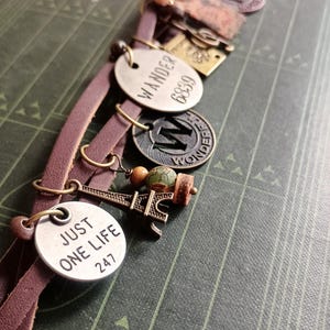 Könnte beinhalten: Ein braunes Lederarmband mit mehreren Metallanhängern. Die Anhänger umfassen einen runden Anhänger mit dem Text "WANDER 6839", einen Anhänger mit dem Buchstaben "W" und dem Wort "WONDER", einen Anhänger mit einem kleinen Eiffelturm-Design und einen runden Anhänger mit dem Text "JUST ONE LIFE 247".