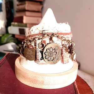 May include: A handcrafted charm bracelet with bronze pendants, including a floral design and the words "live your dream." The bracelet is displayed on a white, cone-shaped stand with a wooden base, showcasing a bohemian style.