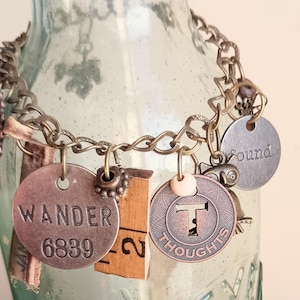 May include: A brass chain bracelet with multiple charms, including a round charm with the words "WANDER 6839", a round charm with the word "THOUGHTS" and a letter "T", a round charm with the word "found", and a small wooden charm with the number "2".