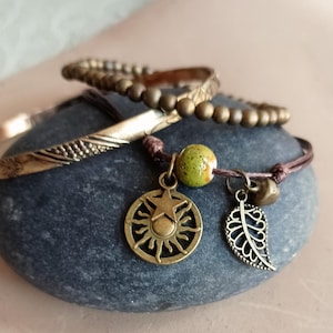 May include: A set of four bracelets with a brown leather cord bracelet with a sun charm, a star charm, a green bead, and a leaf charm. The bracelets are stacked on a gray stone.