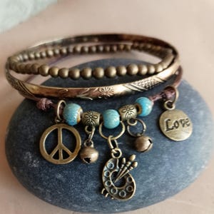 Puede incluir: Un conjunto de pulseras de estilo bohemio. El conjunto incluye varias pulseras de bronce y una pulsera de abalorios con un símbolo de la paz, una paleta de pintor y un dije de 'Amor'. Las pulseras son de color bronce, marrón y turquesa.