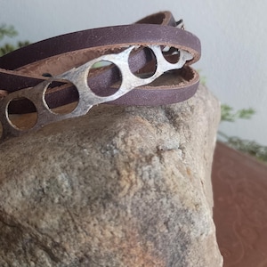 May include: A brown leather wrap bracelet with a silver-tone metal accent. The bracelet is wrapped around a textured rock. The metal accent has circular cutouts. The leather appears to be a dark brown colour.