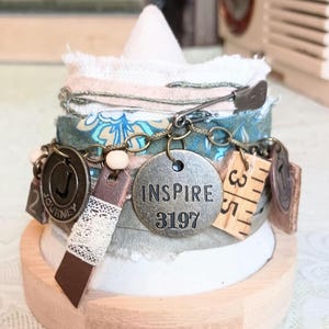 May include: A handcrafted fabric bracelet with various charms. The bracelet features a safety pin, metal tags with the words "INSPIRE 3197" and "JOURNEY", and a small wooden ruler piece. The bracelet is adorned with cream-colored lace and beads.