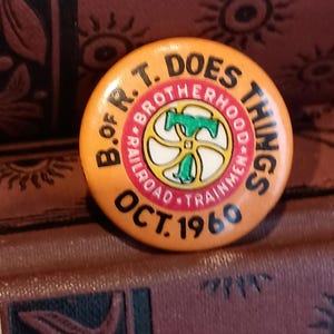 May include: A round, orange button with black text that reads "B. OF R.T. DOES THINGS" and "RAILROAD TRAINMEN" in a circle. The center features a green and white design. The button also has "OCT. 1960" printed on it.