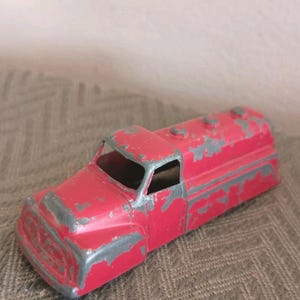 May include: A vintage red toy truck with a tanker body. The truck shows signs of wear, with chipped paint revealing a gray undercoat. The cab has a window, and the tanker has three raised sections on top. A classic collectible.