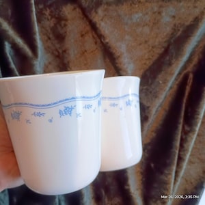 May include: Two white ceramic mugs with a light blue floral pattern around the rim. The mugs have a simple, classic design, suitable for tea or coffee. The mugs are clean and appear to be in good condition.