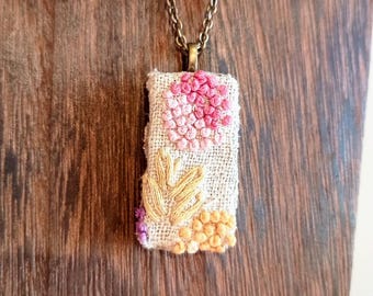 May include: Embroidered pendant necklace with a rectangular pendant. The pendant features a floral design in shades of pink, yellow, and orange. The necklace has a bronze-coloured chain and clasp. The pendant is made of fabric.