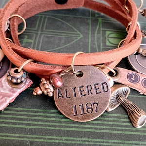 May include: A brown leather wrap bracelet with multiple charms, including a round metal charm with the words "ALTERED 1187" engraved on it, a mushroom charm, and a charm with the word "JOURNEY" on it.
