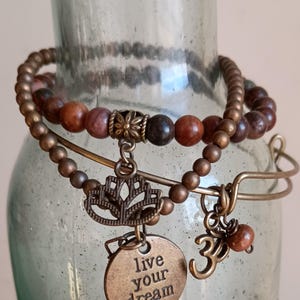 Puede incluir: Pulseras de bronce con abalorios decorativos. Una pulsera tiene cuentas marrones, otra tiene un abalorio de flor de loto y un abalorio redondo con la inscripción "live your dream". Las pulseras están apiladas.