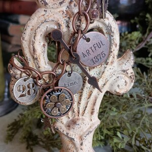 Peut inclure: Un bracelet en chaîne de cuivre avec plusieurs breloques, dont un breloque rond avec le texte "ARTFUL 1375", un breloque rond avec le numéro "28", un breloque rond avec le texte "time.", et un breloque d'engrenage avec un motif de fleur.