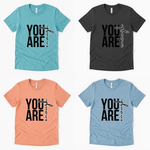May include: Four t-shirts in different colors: teal, charcoal, peach, and light blue. Each shirt has the words "YOU ARE" in bold black letters, with "enough" written in a cursive font.