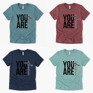 May include: Four short-sleeved t-shirts in teal, burgundy, navy, and seafoam green. Each shirt features the words "YOU ARE" in bold black letters, with the word "enough" written in a cursive font. The shirts are displayed on a white background.