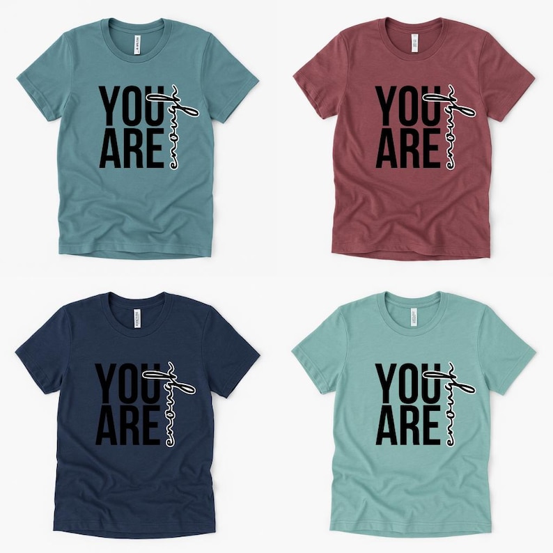 May include: Four short-sleeved t-shirts in teal, burgundy, navy, and seafoam green. Each shirt features the words "YOU ARE" in bold black letters, with the word "enough" written in a cursive font. The shirts are displayed on a white background.
