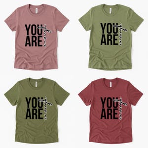 May include: Four short-sleeved t-shirts in various colors: dusty rose, olive green, and burgundy. Each shirt features the phrase "YOU ARE" in bold black letters, with the word "strong" written in a cursive font.