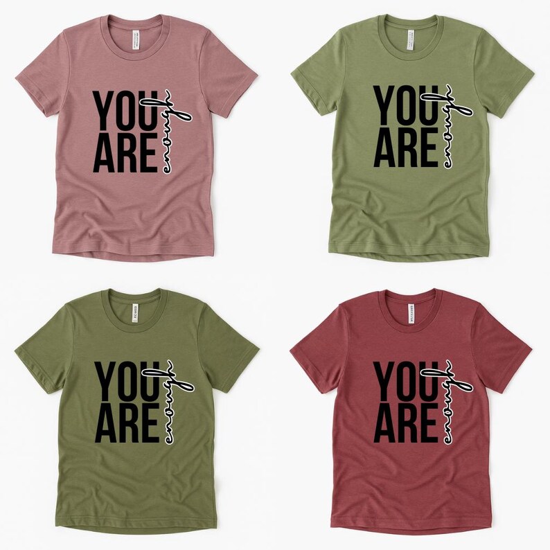 May include: Four short-sleeved t-shirts in various colors: dusty rose, olive green, and burgundy. Each shirt features the phrase "YOU ARE" in bold black letters, with the word "strong" written in a cursive font.