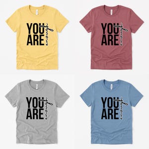 May include: Four short-sleeved t-shirts in yellow, burgundy, gray, and blue. Each shirt features the text "YOU ARE" in a bold, black font, with "enough" written in a script font. The shirts are displayed on a white background.