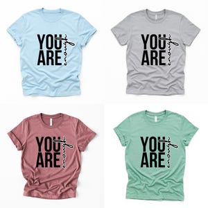May include: Four short-sleeved t-shirts in light blue, gray, mauve, and mint green. Each shirt features the words "YOU ARE" in bold black letters, with "enough" written in a cursive font.