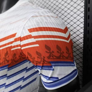 May include: White and orange short-sleeved shirt with a geometric design. The shirt features orange horizontal stripes and a blue and white pattern on the sleeve cuffs. The fabric has a textured appearance.