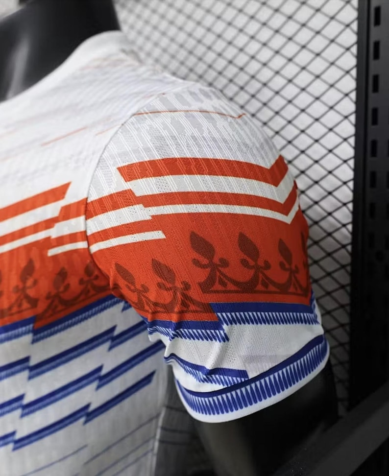 May include: White and orange short-sleeved shirt with a geometric design. The shirt features orange horizontal stripes and a blue and white pattern on the sleeve cuffs. The fabric has a textured appearance.