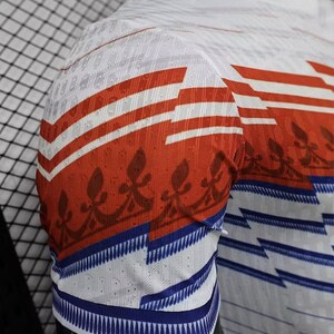 May include: White short-sleeve shirt with red and white horizontal stripes on the sleeves. The sleeves also feature a blue and white striped pattern at the cuff and a brown floral design. The shirt is made of a breathable, textured fabric.