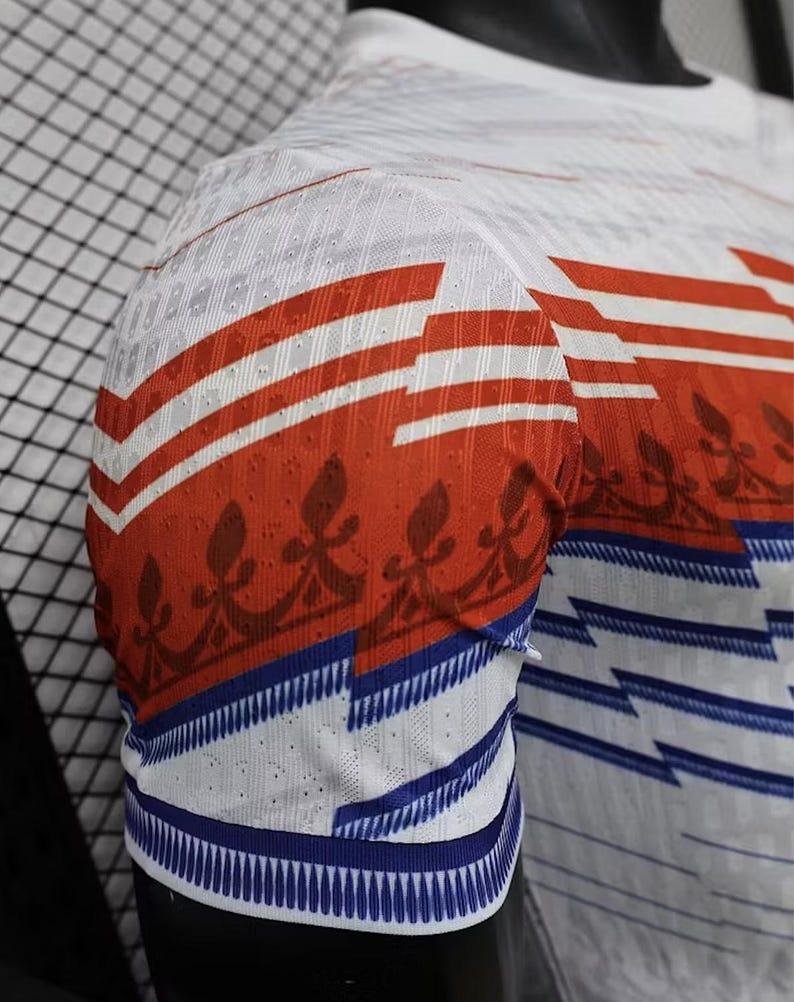 May include: White short-sleeve shirt with red and white horizontal stripes on the sleeves. The sleeves also feature a blue and white striped pattern at the cuff and a brown floral design. The shirt is made of a breathable, textured fabric.